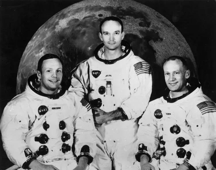 Το πλήρωμα του Apollo 11, Neil Armstrong, Michael Collins και Buzz Aldrin, με τις στολές τους μπροστά σε φωτογραφία της Σελήνης